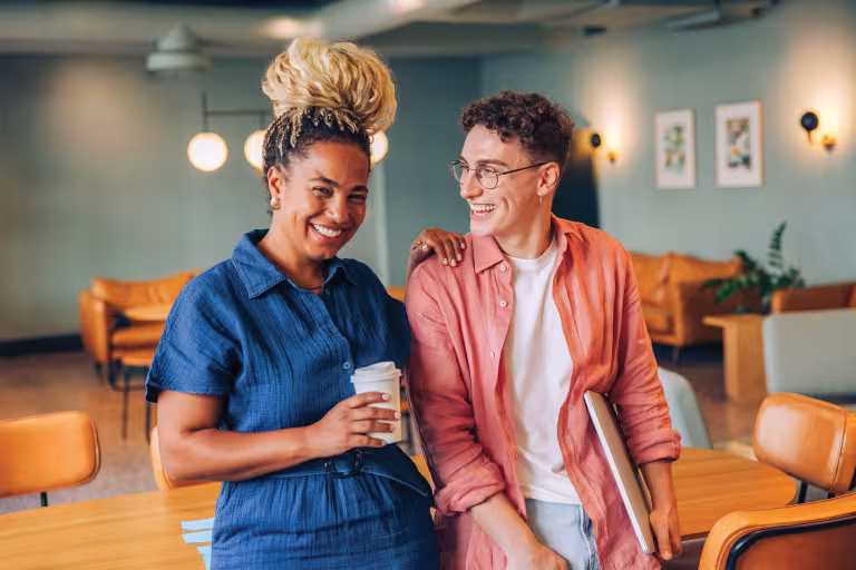Two young adults meet up in a coffee shop and have a casual conversation, the person on the left is holding a coffee cup and is smiling at the camera. 