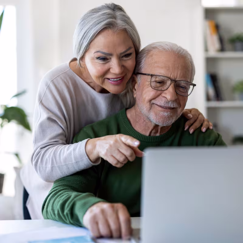 A retired couple is using a computer, the woman standing over the mans shoulder points at the screen. 