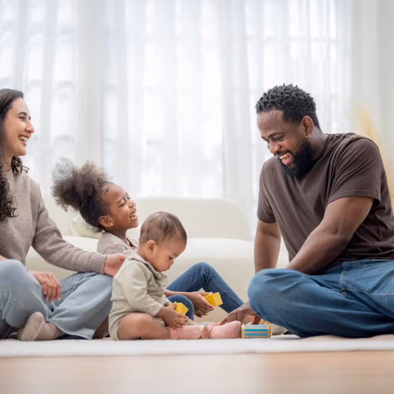 A young family with two parents and two children sit on the floor laughing and playing with toys. 
