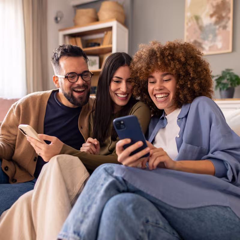 Three friends in their thirties sit together on a couch looking at something on their phones