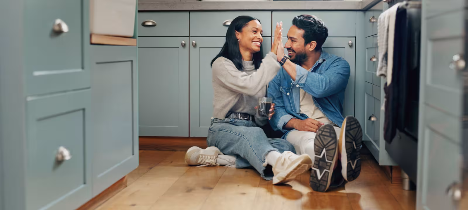 A young couple sits on the floor of their kitchen drinking coffee and sharing a high five 