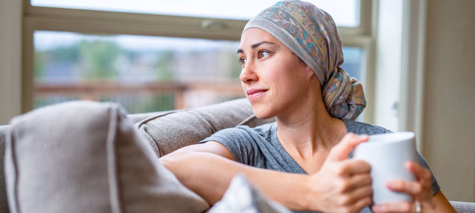A woman with cancer wears a headscarf after losing her hair and sits on her couch with a mug of tea while looking out a window