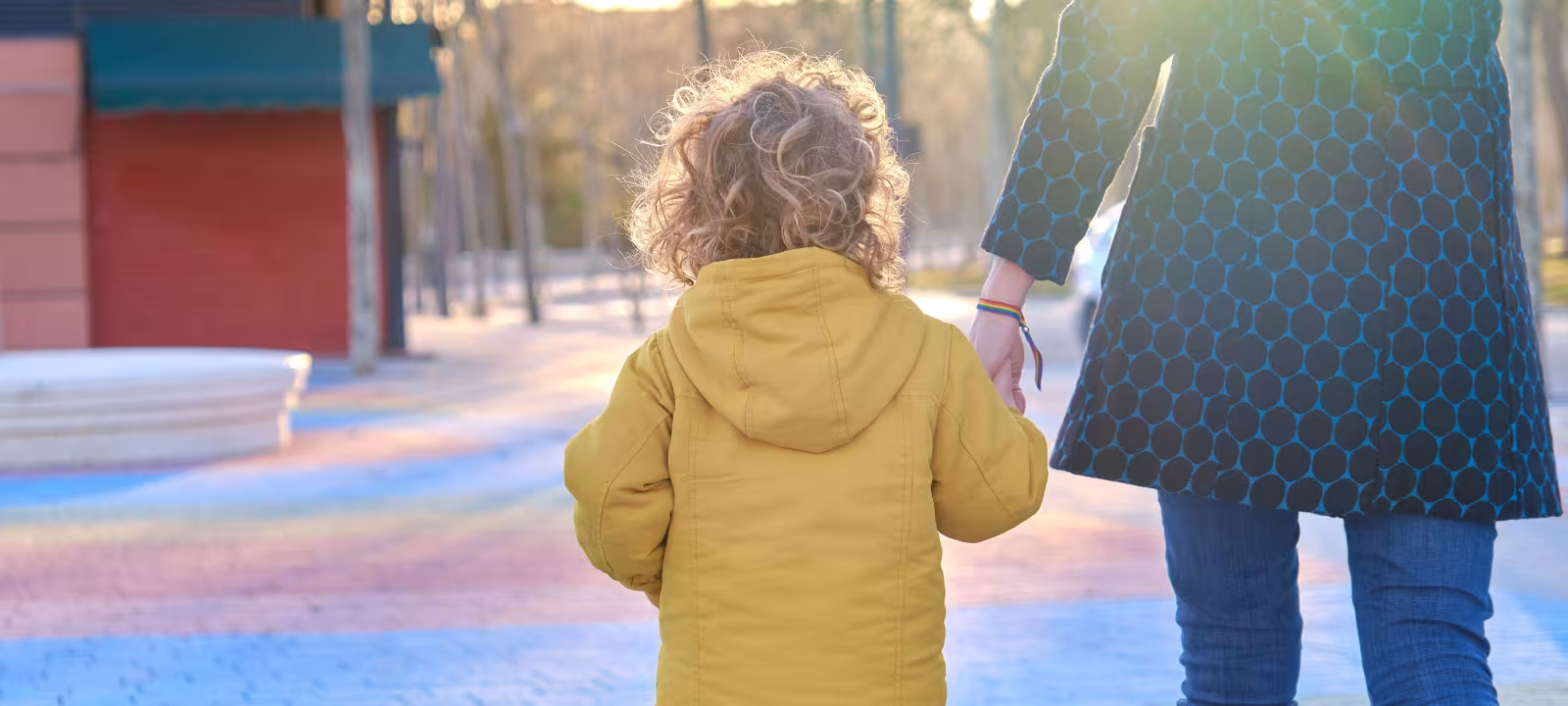 A young child holds an adults hand as they walk away from the camera through a park