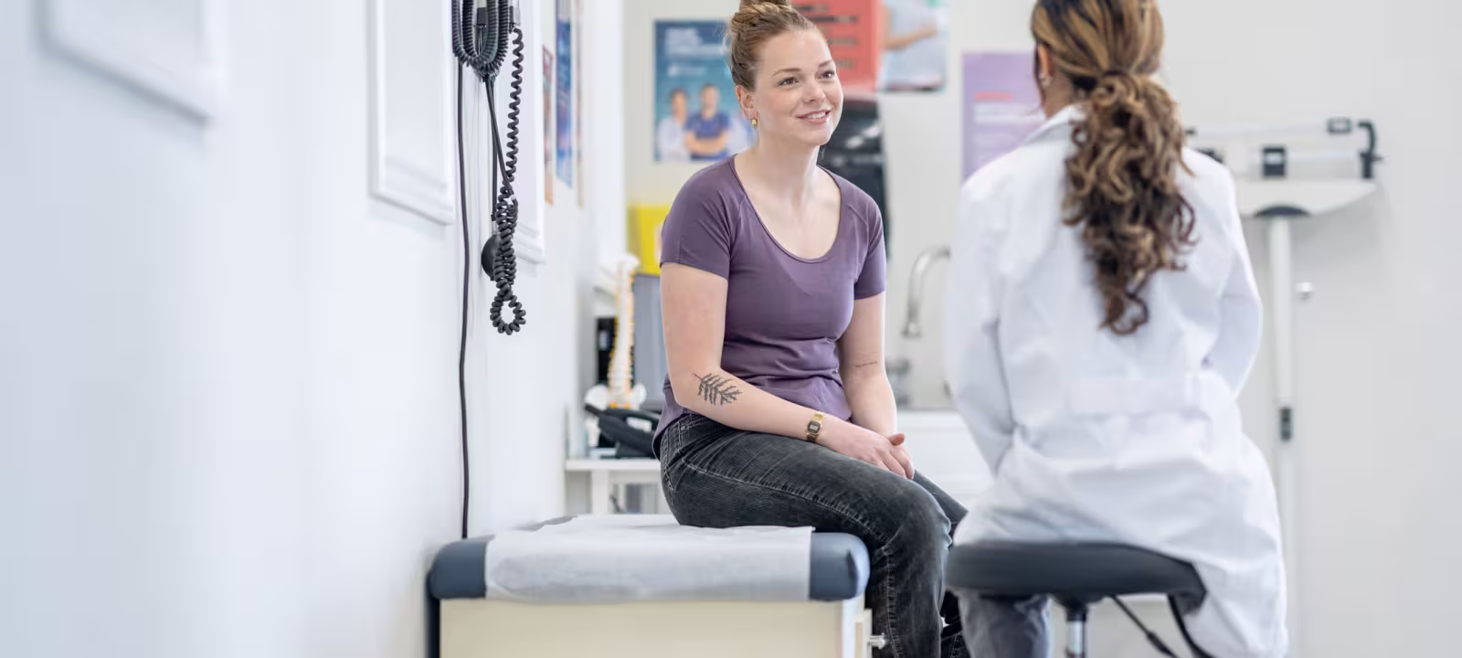 A younger female patient sits on an exam bench while talking to her doctor 