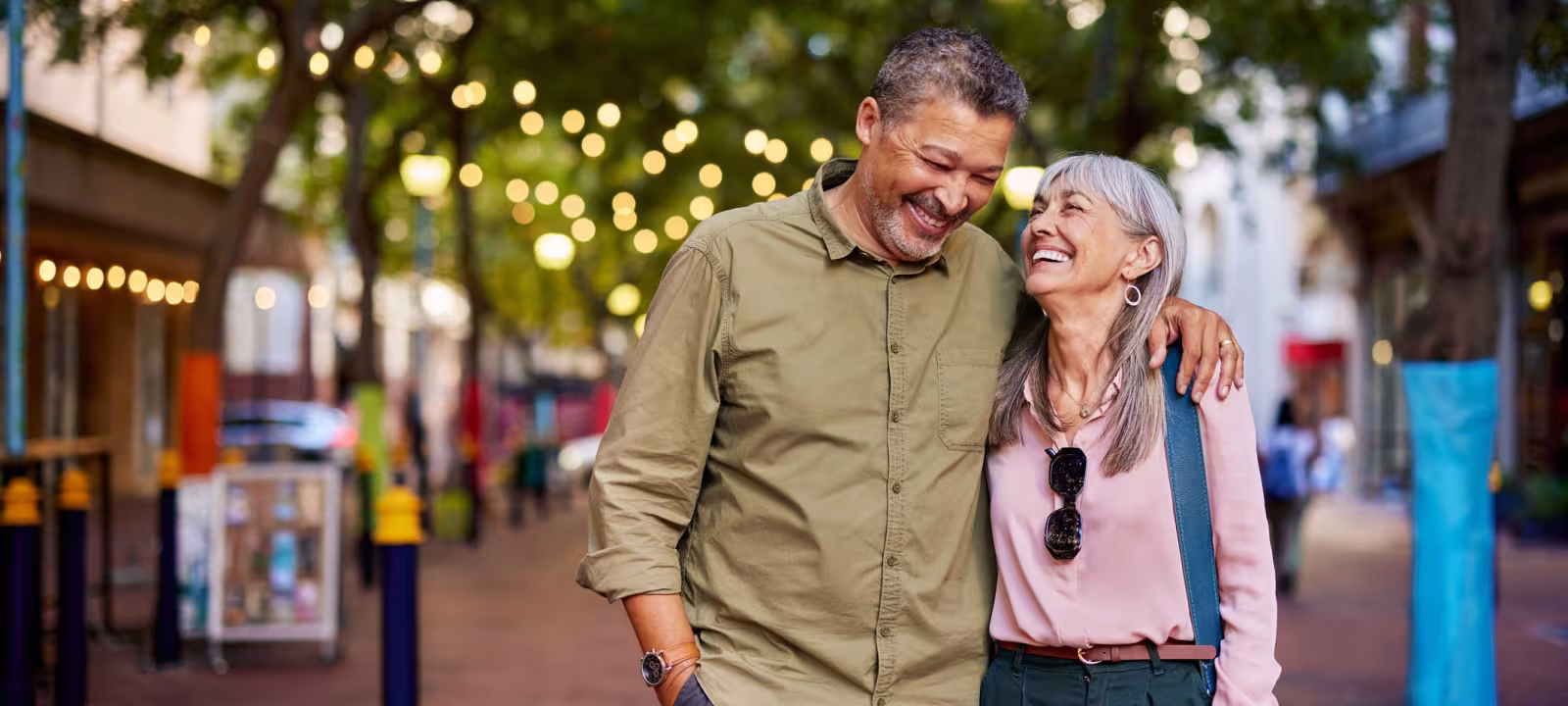 An older couple walks down a pedestrian street lit with twinkle lights as they hug and smile at each other.