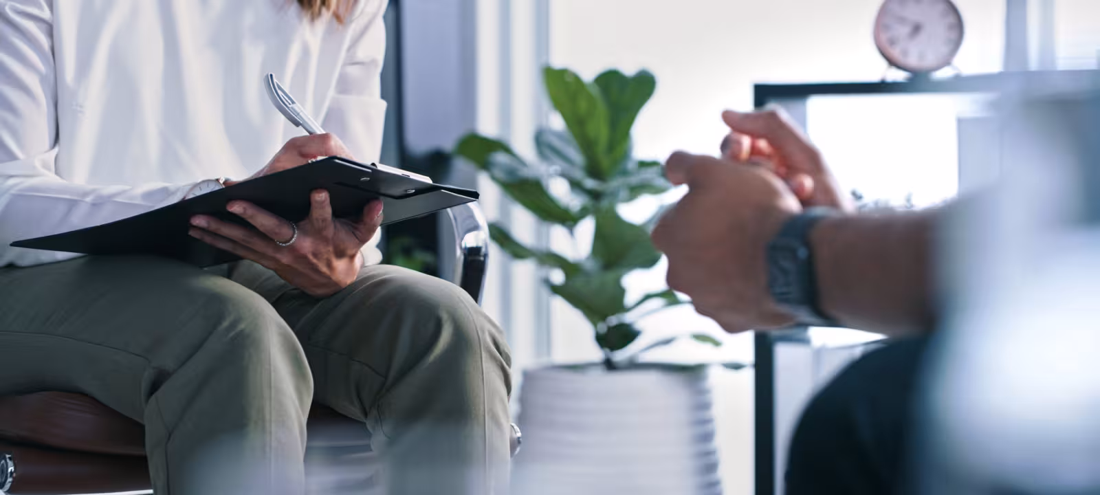 A patient sits across from a therapist who is taking notes on a clipboard, neither persons face is in frame as the image is shot from lower and you can only see them seated. 