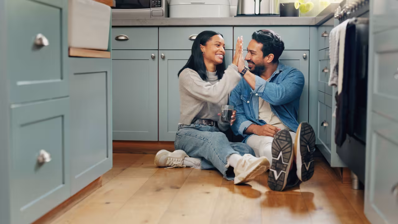 A young couple sits on the floor of their kitchen drinking coffee and sharing a high five 