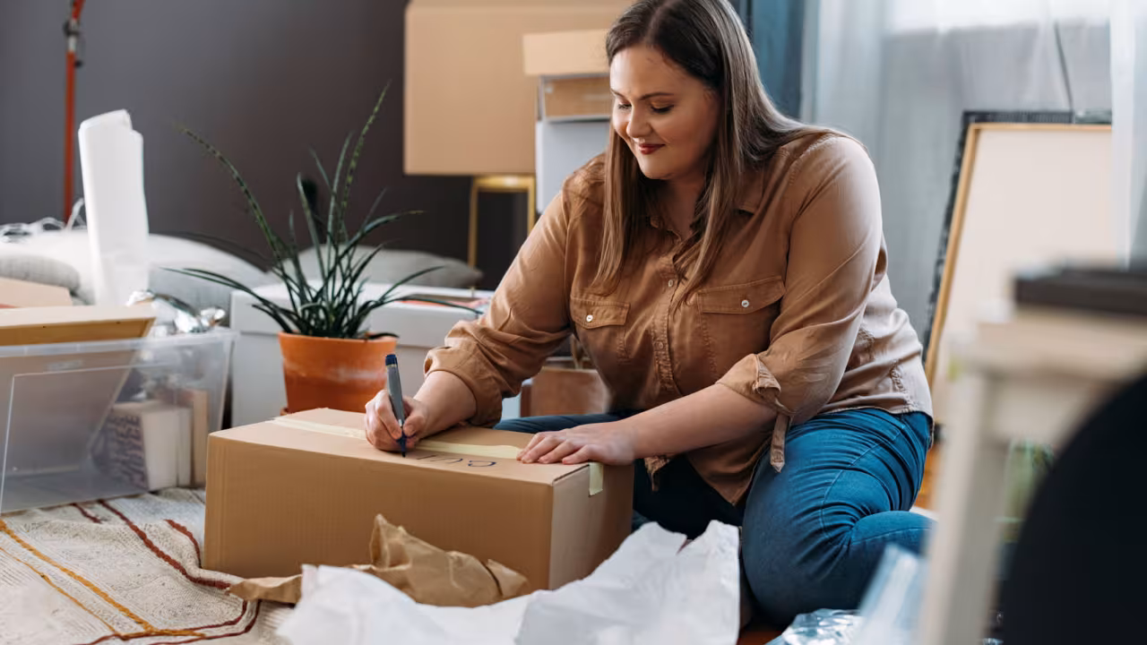 Woman sits on her floor surrounded by packing boxes as she labels one box with a sharpie ahead of a move. 