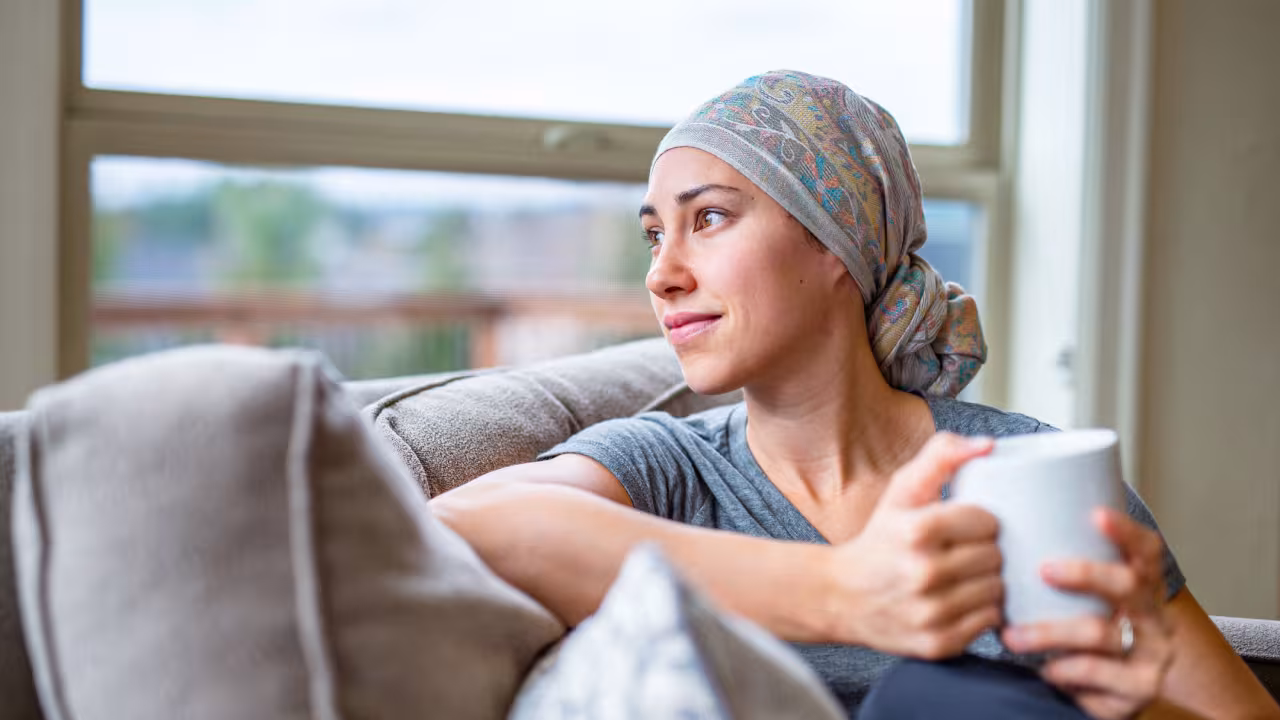 A woman with cancer wears a headscarf after losing her hair and sits on her couch with a mug of tea while looking out a window
