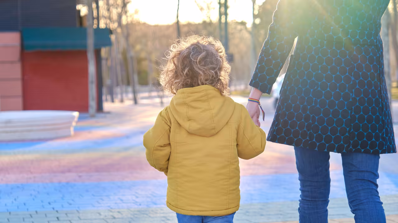 A young child holds an adults hand as they walk away from the camera through a park