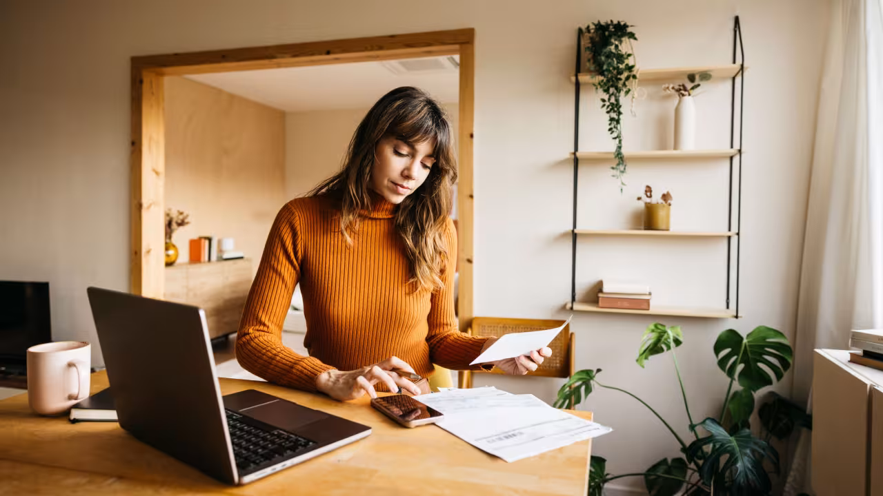 A woman sits at her desk reviewing paperwork and calculating a bill. 