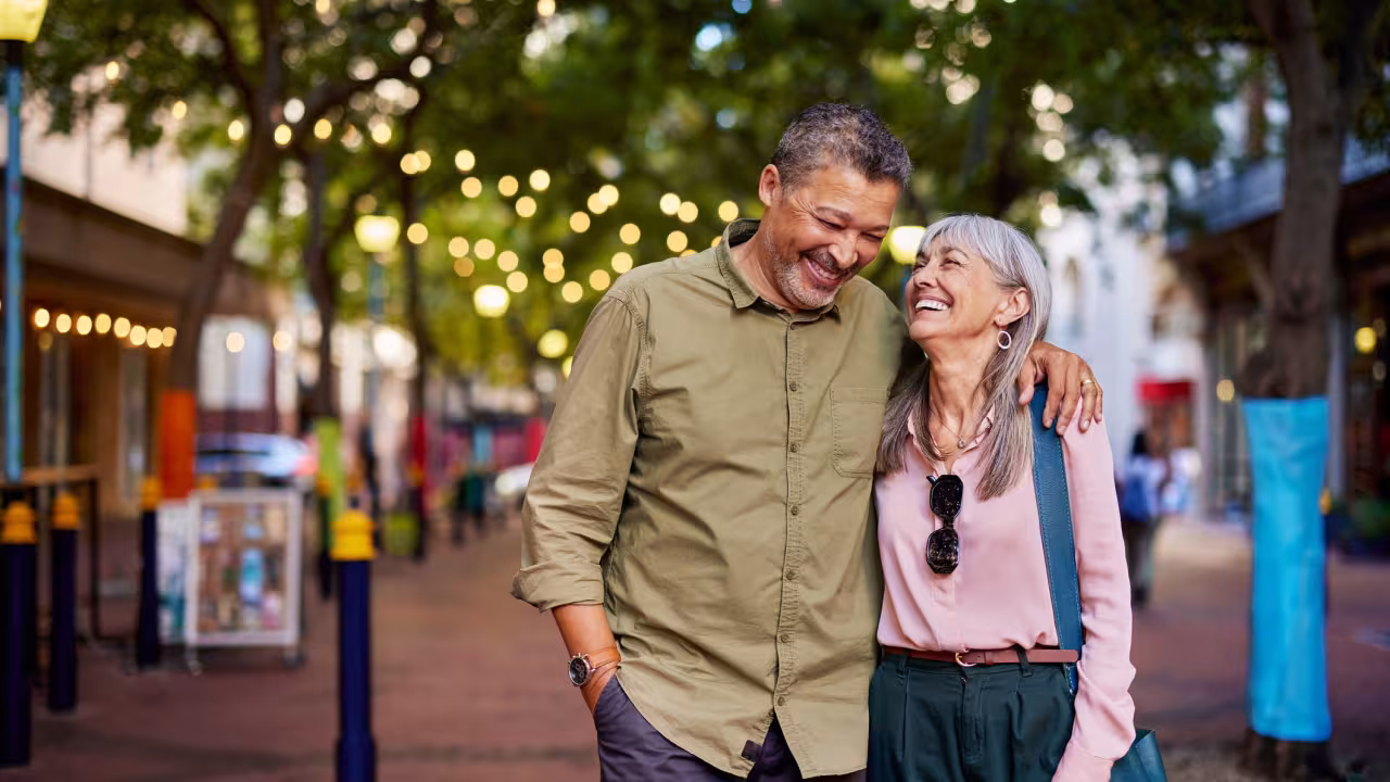 An older couple walks down a pedestrian street lit with twinkle lights as they hug and smile at each other.