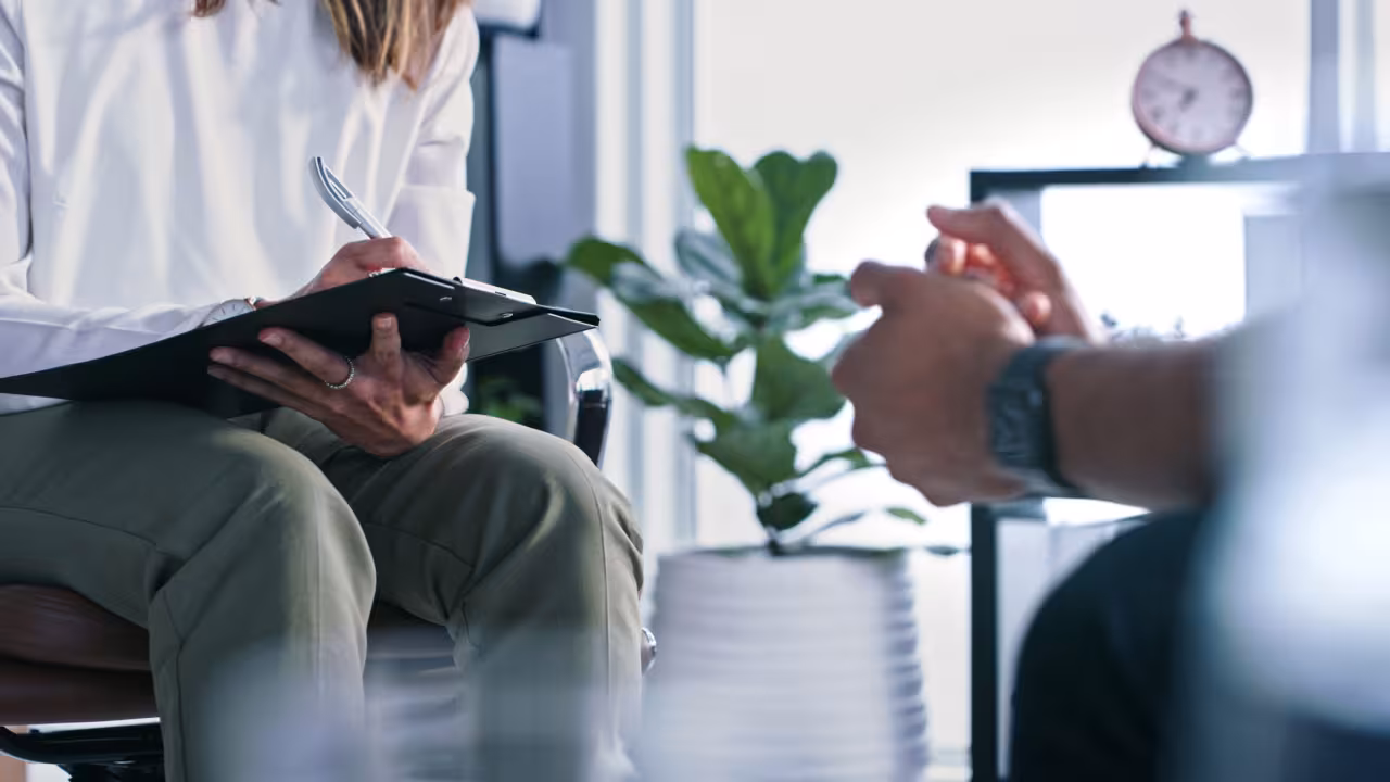 A patient sits across from a therapist who is taking notes on a clipboard, neither persons face is in frame as the image is shot from lower and you can only see them seated. 